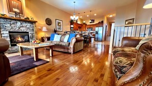 A living room with a fireplace, wooden flooring, leather sofas, a dining area, and a kitchen in the background.