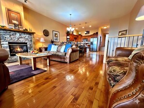 A living room with a fireplace, wooden flooring, leather sofas, a dining area, and a kitchen in the background.