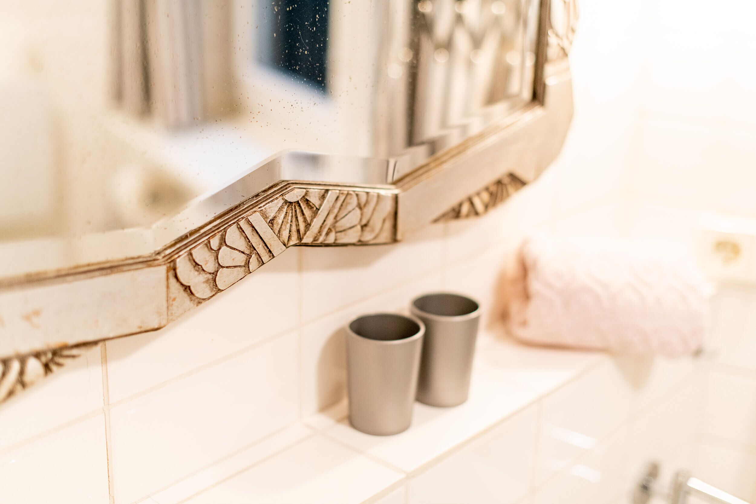A bathroom with a mirror, two grey cups on a shelf, and a pink towel on the floor.