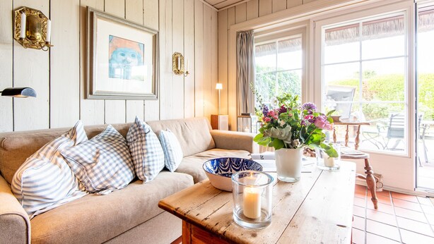 Een gezellige woonkamer met een houten vloer, een beige bank met decoratieve kussens, een houten salontafel, een lijst met een afbeelding aan de muur, en een raam met gordijnen.