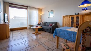 Ein Wohnzimmer mit einer Couch, einem kleinen runden Tisch, einem Esstisch mit blauer Tischdecke und einem Schrank.