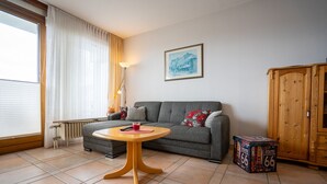 Ein Wohnzimmer mit grauem Sofa, einem runden Holztisch, einer Heizung, einer Lampe, einem Schrank und einem gerahmten Bild an der Wand.