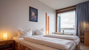 Ein Hotelzimmer mit zwei Betten, einem Nachttisch, einer Lampe, einem Fenster mit Vorhängen, einem Heizkörper und einem gerahmten Bild an der Wand.