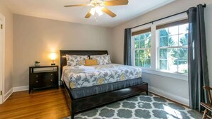 Une chambre à coucher comprenant un lit, une table de chevet, un ventilateur de plafond, une fenêtre avec des rideaux et un tapis à motifs.