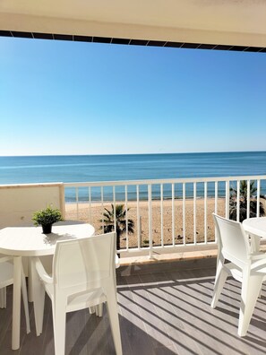 A balcony with white chairs and a small table, overlooking a beach with a clear view of the ocean.