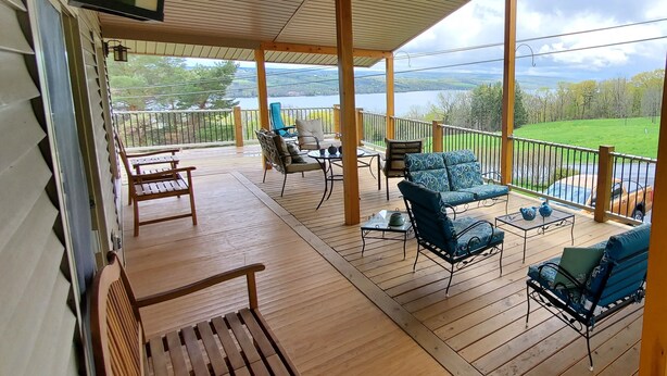A covered outdoor deck with wooden flooring, metal railings, and furniture including chairs and a table. - Hillside Haven (Penn Yan)