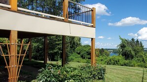 A two-story wooden deck with a metal railing, supported by wooden pillars, overlooking a well-maintained lawn and trees. - Hillside Haven (Penn Yan)