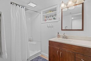 A modern bathroom with a walk-in shower, a vanity with a mirror, and a shelf with toiletries. - Sweet & Cozy Studio Apartment (South Burlington)