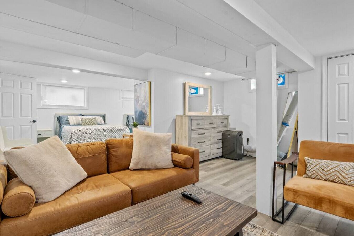 Ein modernes, helles Zimmer mit einem braunen Ledersofa, einem Holztisch und einem kleinen Sessel. Im Hintergrund befindet sich ein Bett mit gemustertem Bettzeug, eine Kommode mit Spiegel und ein Bild an der Wand.