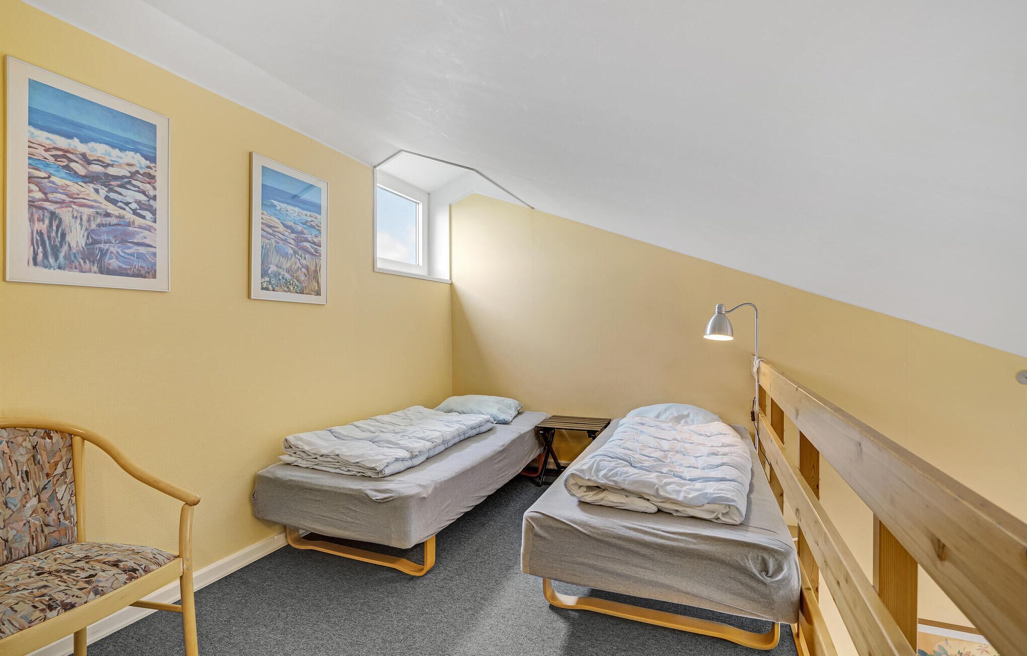 A small, cozy room with two single beds, a chair, and two paintings on the wall.