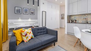 Uma sala de estar moderna com um sofá cinzento, um travesseiro amarelo e um travesseiro colorido. Há três quadros nas paredes, um armário branco e uma área de cozinha com painel de azulejos.