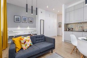 Una sala moderna con un sofá gris, un cojín amarillo y un cojín colorido. Hay tres cuadros enmarcados en la pared, un mueble blanco y una zona de cocina con salpicadero de azulejos.