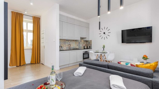Uma sala de estar moderna com um sofá cinza, uma mesa de jantar com uma bandeja de comida, uma TV fixada na parede e uma cozinha ao fundo.