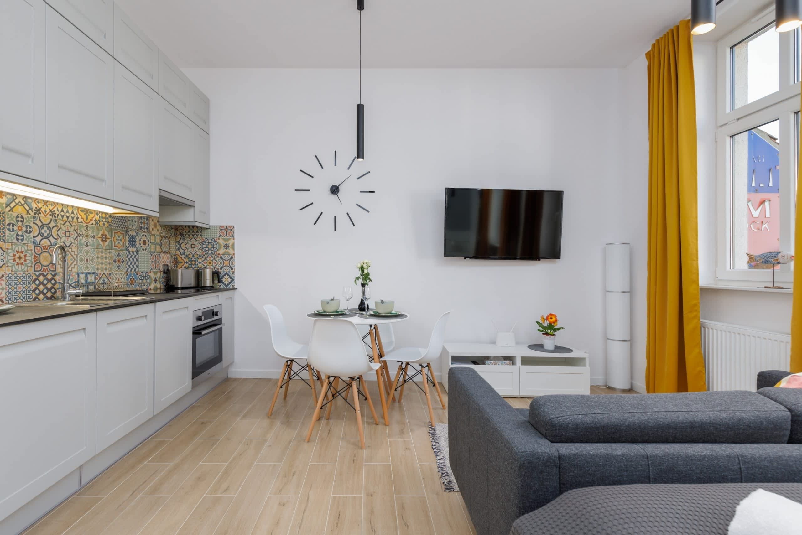 A modern kitchen with white cabinets, a tiled backsplash, a dining area with white chairs, a gray sofa, and a flat-screen TV mounted on the wall.