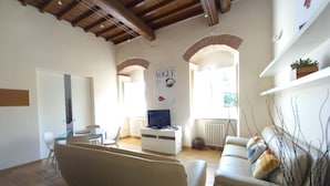 A spacious living room with a high ceiling, a white sofa, a television, and a window with a view.