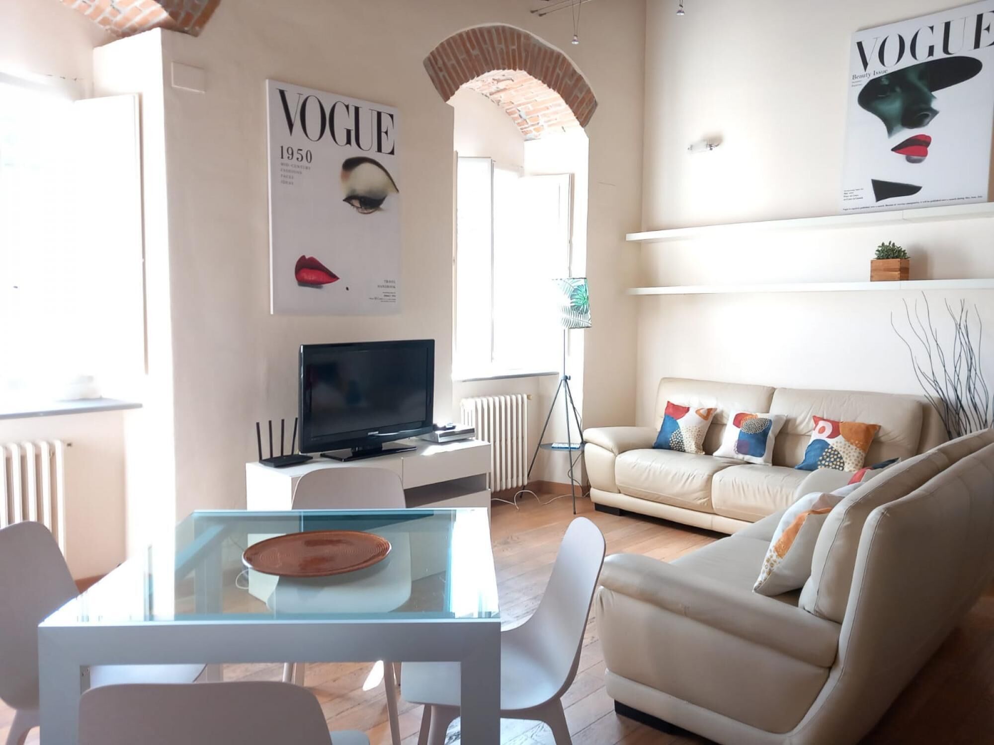 Una sala de estar moderna con una mesa de comedor de vidrio, un televisor de pantalla plana, un sofá color beige y una revista en la pared.