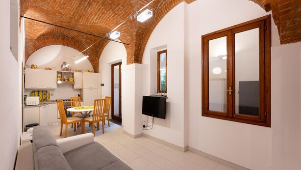 A modern living room with a grey sofa, wooden chairs, a TV, and a kitchen in the background. - San Zanobi 33 in Firenze (Florence)