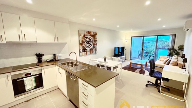 A modern kitchen with a central island, a dining area with a white table and chairs, a living area with a sofa and a TV, and a large sliding glass door leading to a balcony. - Beige & Beautiful - Close to ANU (Turner)