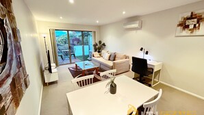 A modern living room with a white sofa, a dining table, and a TV. There is a balcony with a view of greenery and buildings. - Beige & Beautiful - Close to ANU (Turner)