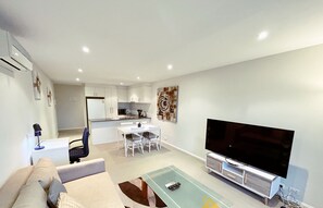 A modern living room with a sofa, a glass coffee table, a TV, and a dining area with chairs and a kitchen in the background. - Beige & Beautiful - Close to ANU (Turner)