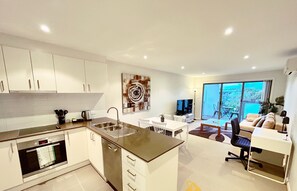 Een moderne keuken met een centraal eiland, een eethoek met een witte tafel en stoelen, een zithoek met een bank en een televisie, en een grote schuifpui naar een balkon.