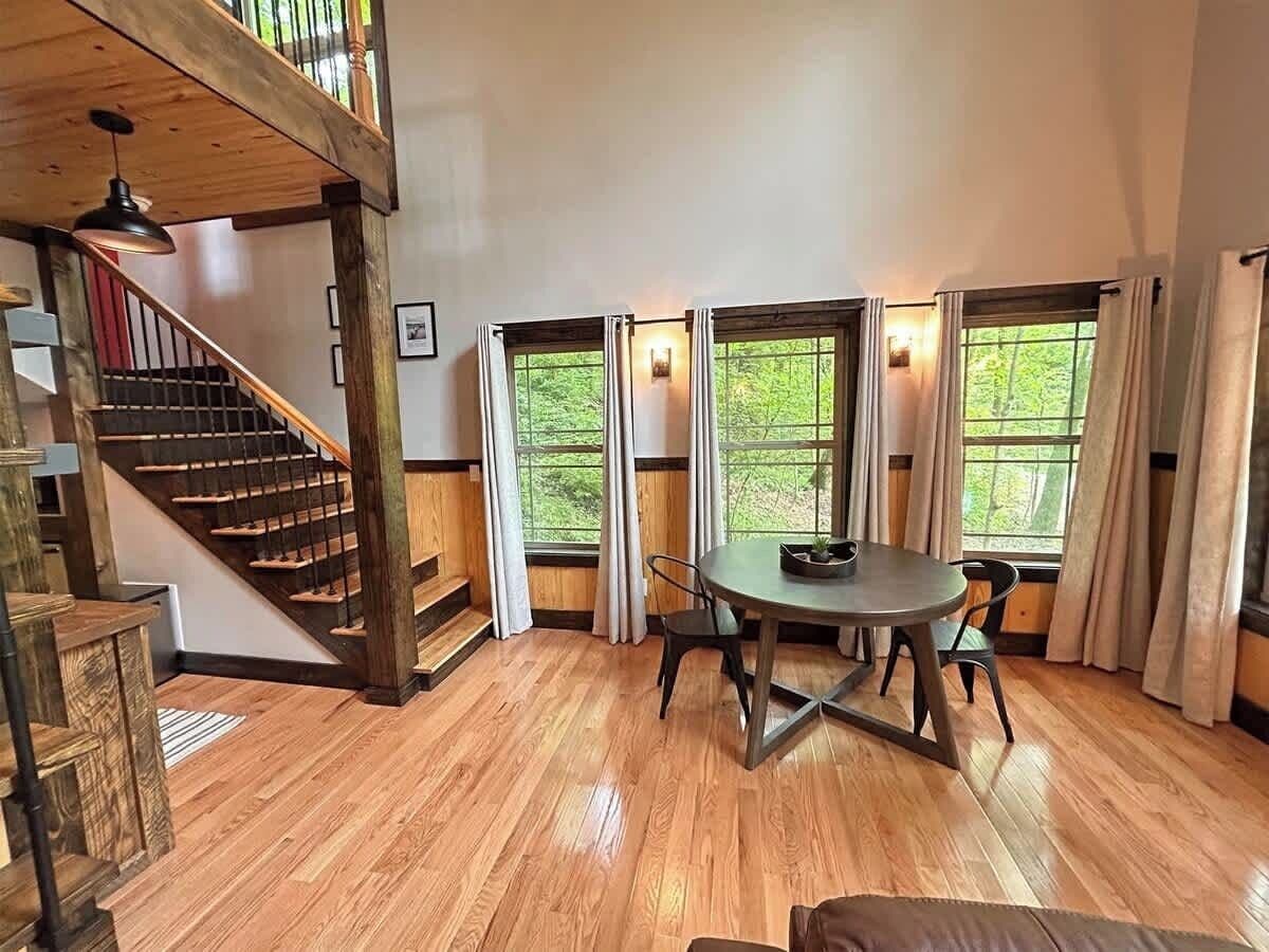 A spacious room with a wooden staircase, a dining table with chairs, and large windows with curtains.