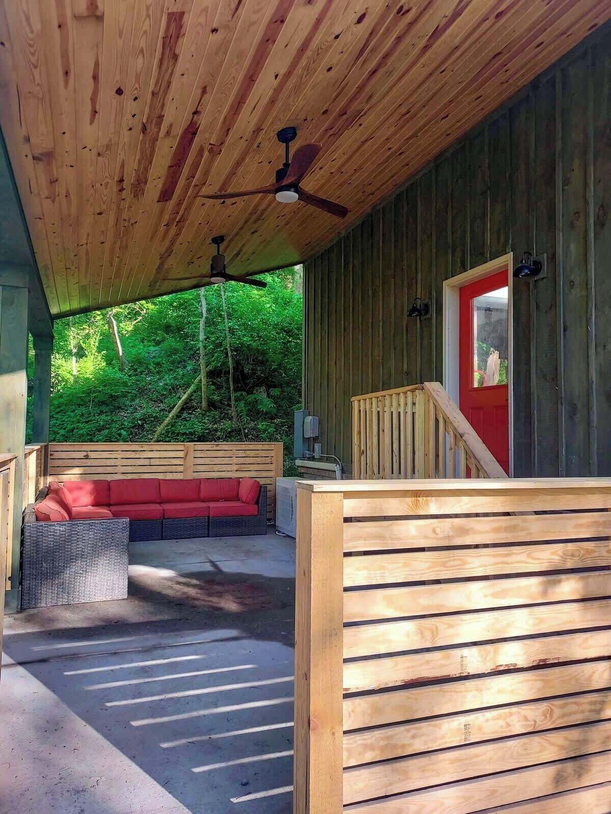 A covered outdoor seating area with a ceiling fan, a red door, and a view of the surrounding greenery.