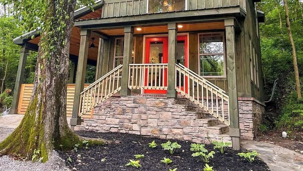 Eine rustikale Holzhütte mit Veranda, Steinstufen und einer roter Tür, umgeben von Bäumen.