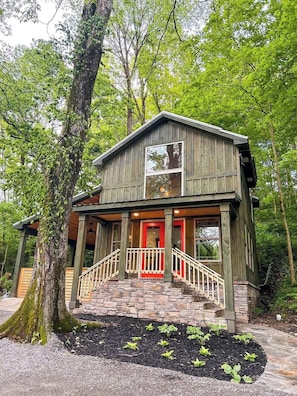 A rustic cabin with a porch, stone steps, and a red door, surrounded by trees.