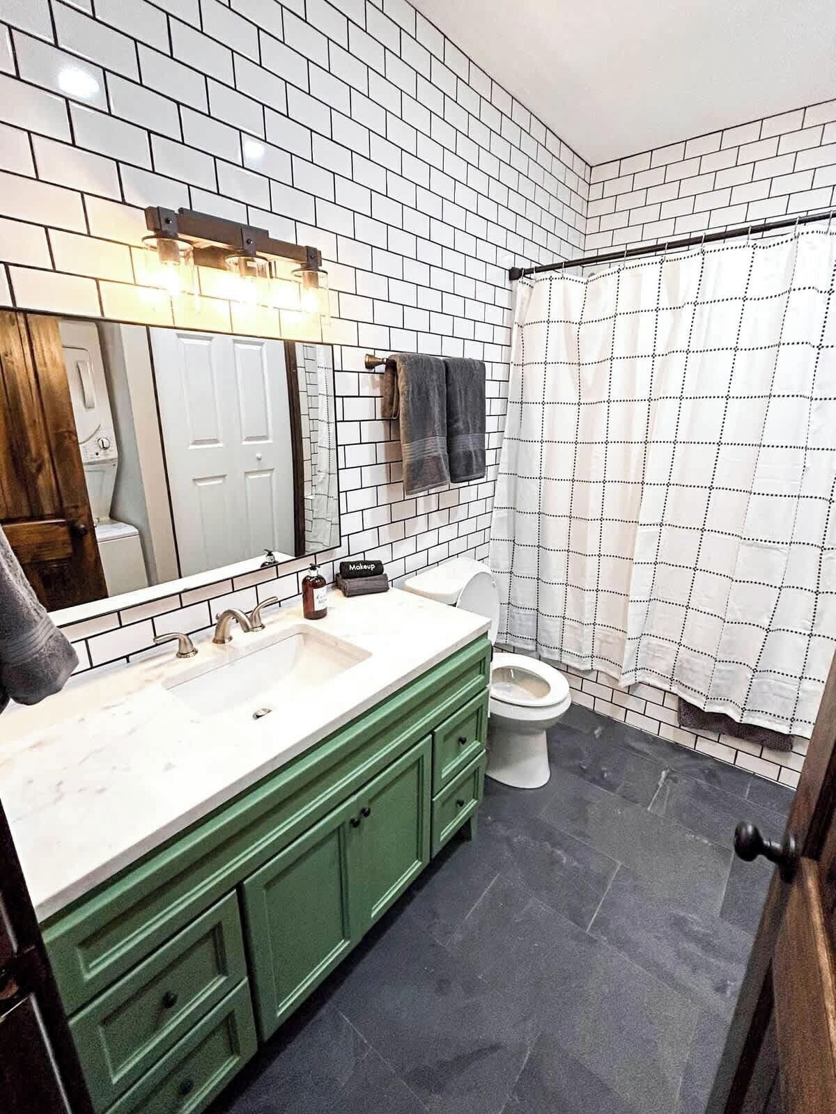 Un baño con pared de azulejos blancos, tocador verde, inodoro blanco y cortina de ducha.