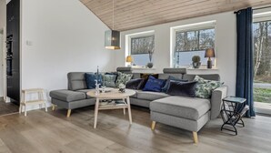 Un soggiorno moderno con un divano angolare grigio, un tavolino rotondo e un pavimento in legno. Si vedono due finestre con tende, una lampada sul davanzale e un piccolo tavolino laterale.