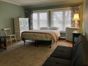 Classic bedroom with a neatly made bed, sofa, and table with a lamp, set amidst a window with blinds and a dresser.
