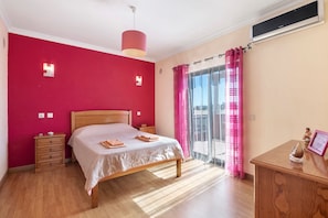 Une chambre à coucher avec un lit, des tables de chevet en bois, une commode, un ventilateur de plafond et une porte-fenêtre coulissante avec des rideaux roses.