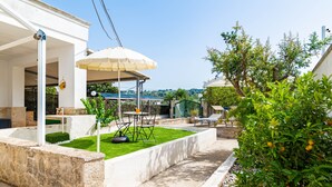 Eine Terrasse mit Tisch und Stühlen, eine überdachte Sitzgelegenheit und ein Garten mit Bäumen und Rasen.
