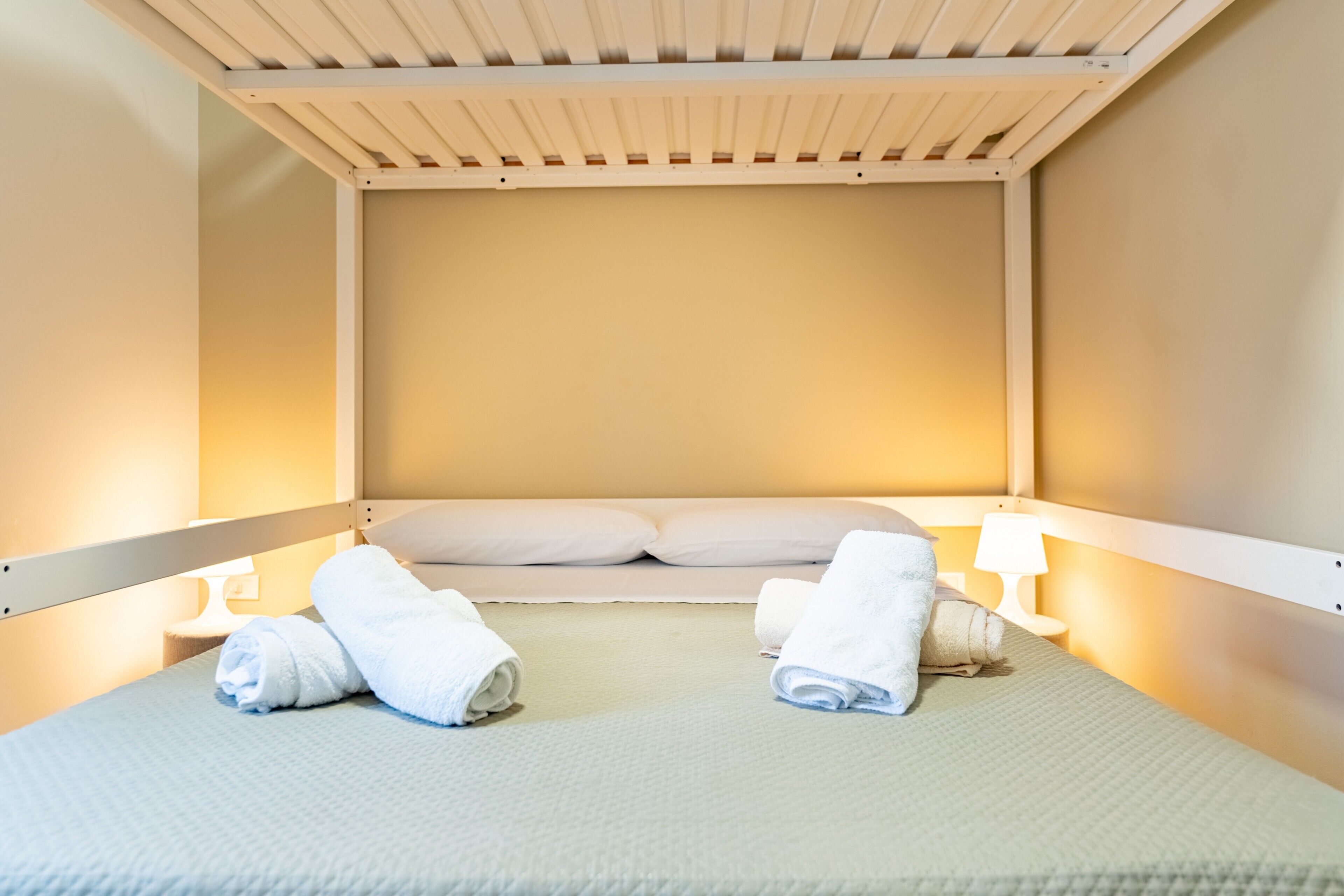 A single bed with white linens, two rolled towels, a nightstand with a lamp, and a wooden ceiling structure.