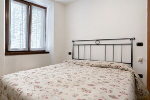 Ein Einzelbett mit Metallgestell, ein Fenster mit Vorhängen und eine schlichte Wand.