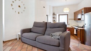 Een woonkamer met een grijze bank, een wandklok, een keuken met houten kasten en een betegelde vloer.