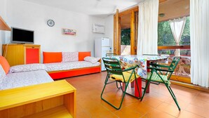 Ein Zimmer mit einer Couch, einem Fernseher, einem Tisch mit Stühlen, einem Deckenventilator und einem Fenster mit Vorhängen.