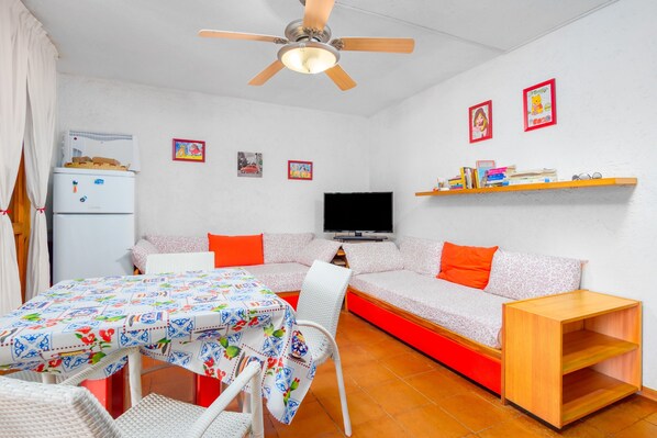 A compact living space with a sofa, a television, a table with a floral tablecloth, a ceiling fan, and a small refrigerator.