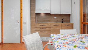 Une cuisine de taille réduite avec des meubles blancs, un plan de travail en bois et un évier. On y trouve une table à manger recouverte d’une nappe colorée, ainsi que deux chaises blanches.