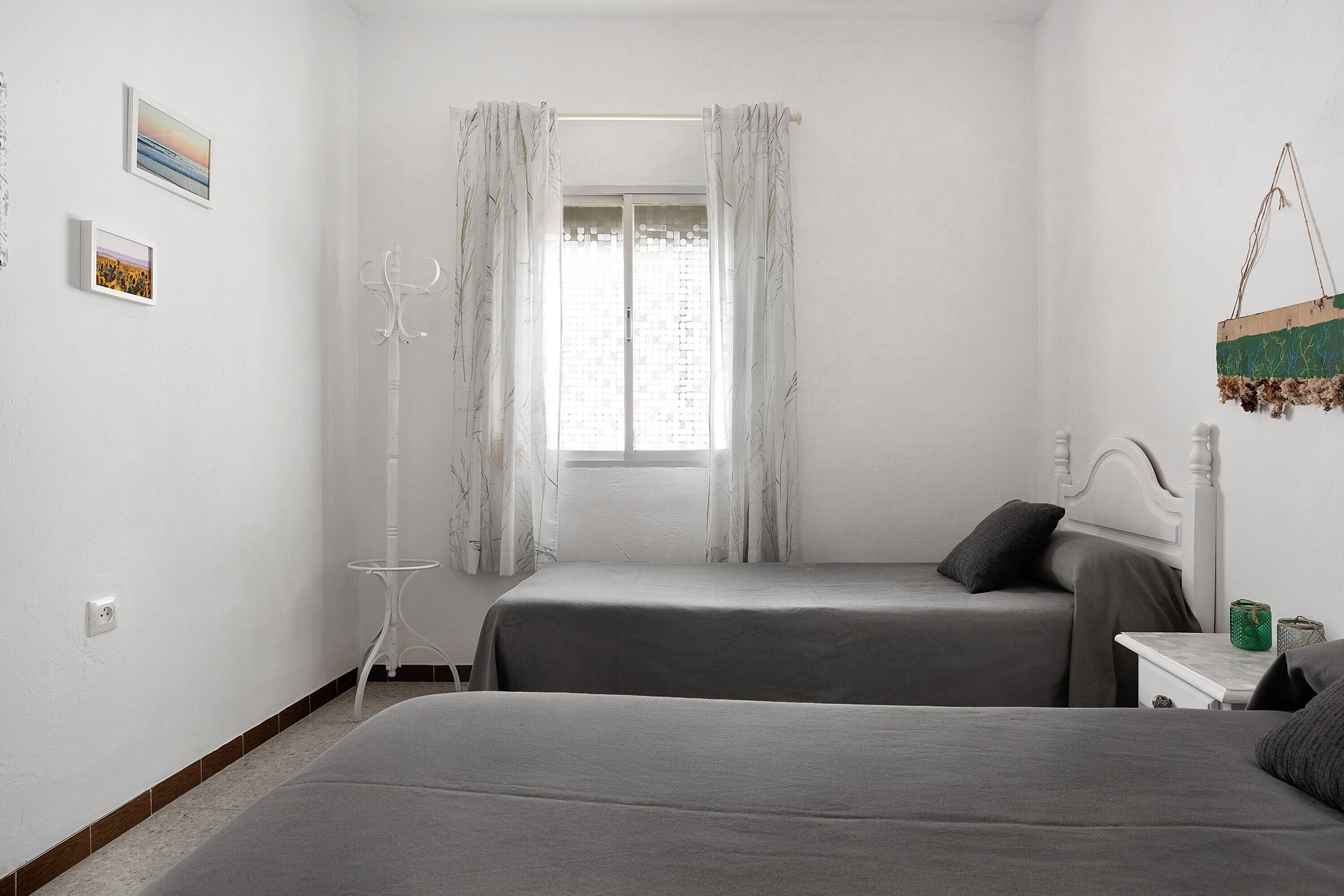 A bedroom with two beds, a window with curtains, a small table, and a wall-mounted hook.