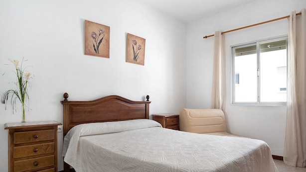 Ein Schlafzimmer mit einem hölzernen Bett, einem Nachttisch, einem Sessel und zwei an der Wand befestigten Bildern.