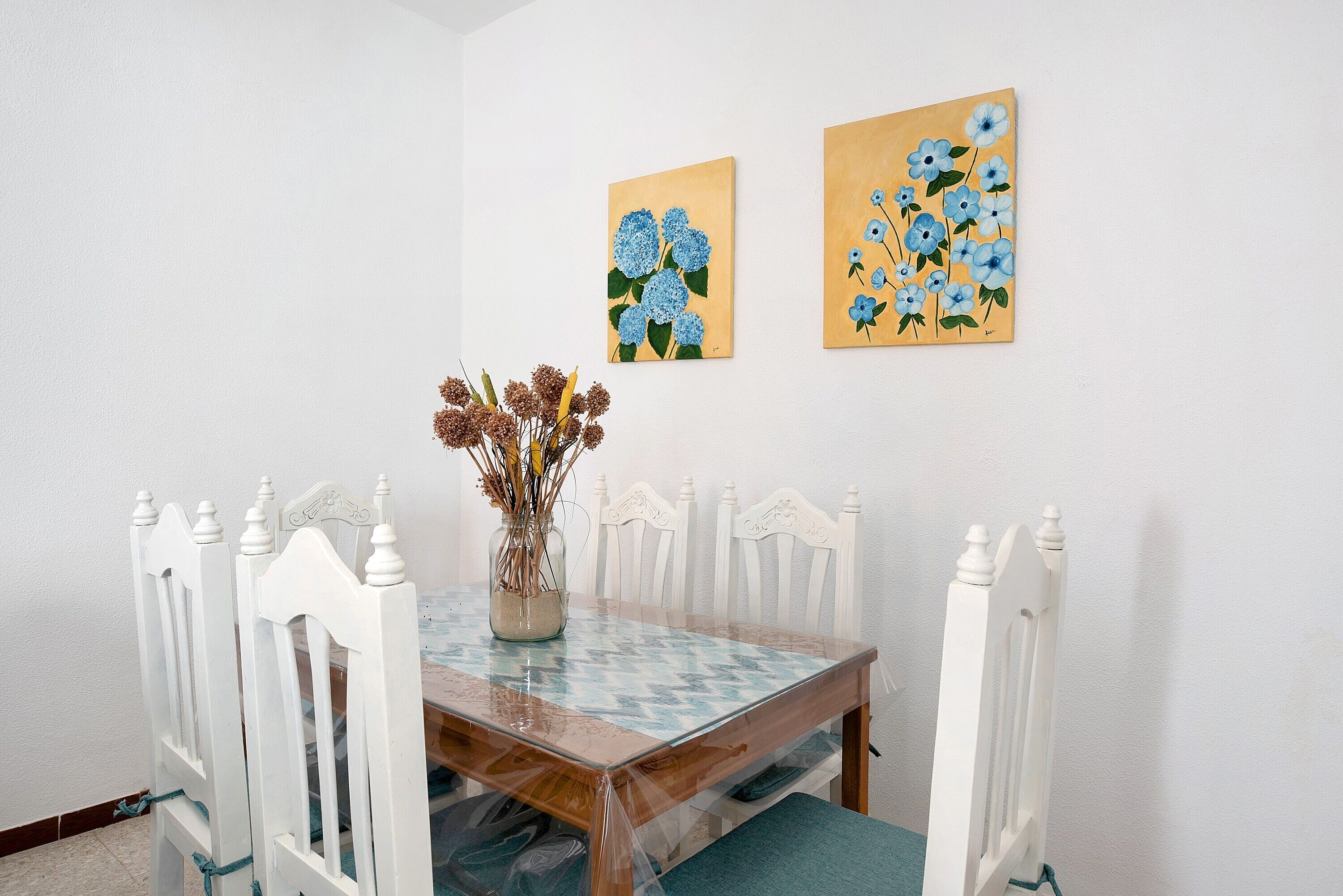 Et spisebord med hvide stole, en vase med tørrede blomster og to billeder på væggen.
