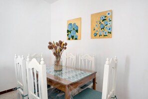 Une table à manger entourée de chaises blanches, un vase avec des fleurs séchées et deux tableaux accrochés au mur.