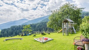Ein Spielplatz mit Rutsche und Klettergerüsten, umgeben von einer Rasenfläche mit Bank und Baum.