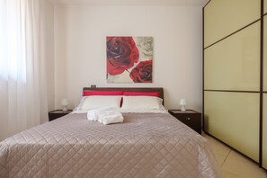 Una cama bien hecha con cubrecama acolchado, almohada roja y blanca, y una obra de arte en la pared con rosas rojas.
