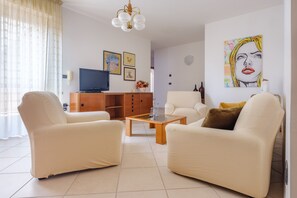 Una sala de estar con dos sofás blancos, una mesa de centro de madera, un televisor sobre un mueble de madera y obras de arte en las paredes.