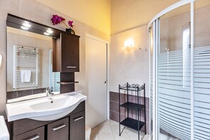 Un baño con un lavamanos blanco, armarios oscuros, cabina de ducha de vidrio y estante metálico negro.