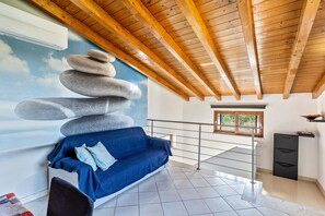 Une pièce avec un canapé bleu, un mur en pierres apparentes, un plafond en bois, une fenêtre et une rambarde en métal.