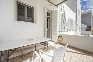 A balcony with a white table and chairs, a window with blinds, and a view of a building.